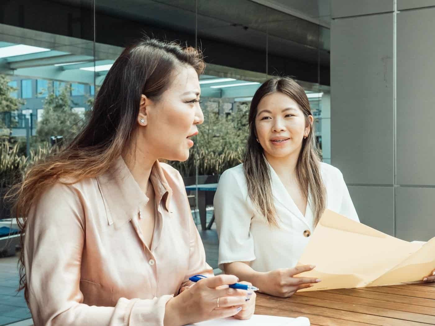 Two Asian businesswomen reviewing paperwork together outside an office building
