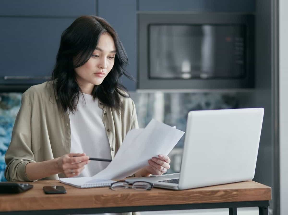 Asian woman reviewing documents beside a laptop while working from home, representing a flexi-place FWA arrangement